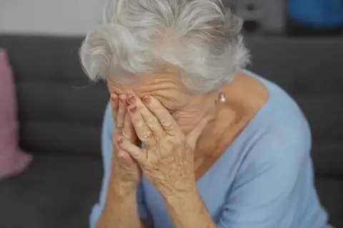 Starsza kobieta z siwymi włosami zakrywa twarz dłońmi, być może doświadczając trudnych emocji związanych z objawami alzheimera.