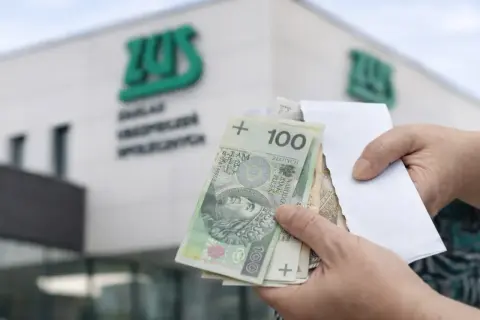 Ręce trzymają polskie banknoty przed budynkiem ZUS. Wypłata 14 emerytury wreszcie nadeszła.