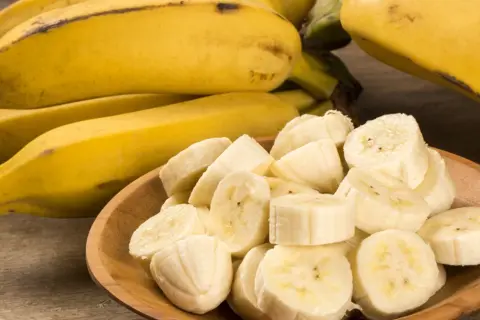 Kawałki dojrzałego banana w drewnianej miseczce, obok całe owoce.