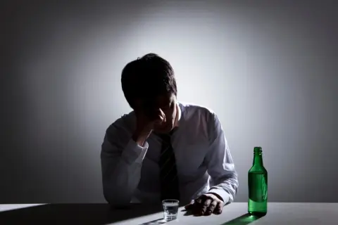 Mężczyzna w białej koszuli i krawacie, z ręką na czole, siedzi przy stole. Obok niego stoi zielona butelka i szklanka. Wygląda na to, że zmaga się z zatruciem alkoholowym.