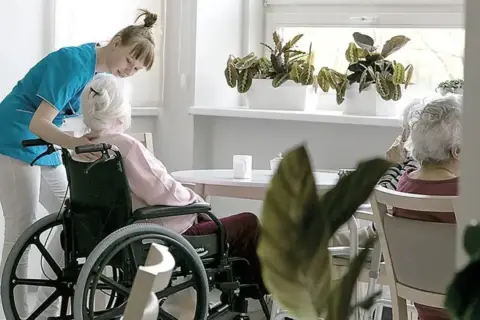 Opiekunka pomaga starszej kobiecie na wózku inwalidzkim. To przykład troski w geriatrii, która zajmuje się zdrowiem seniorów.