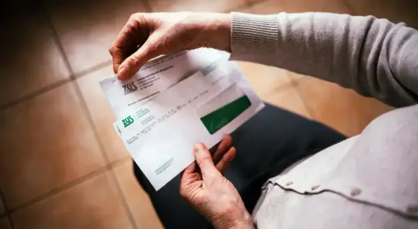 Dłoń otwiera kopertę ZUS. W środku dokumenty dotyczące 13. emerytury, być może decyzja o jej przyznaniu.