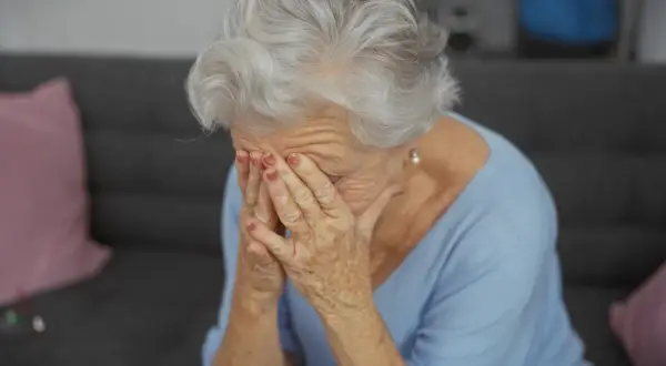 Starsza kobieta z siwymi włosami zakrywa twarz dłońmi, być może doświadczając trudnych emocji związanych z objawami alzheimera.