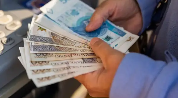 Dłonie trzymają stos polskich banknotów, symbolizujących waloryzację emerytury. Widać nominały 50, 100 i 200 zł.