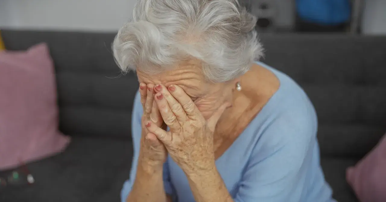 Starsza kobieta z siwymi włosami zakrywa twarz dłońmi, być może doświadczając trudnych emocji związanych z objawami alzheimera.