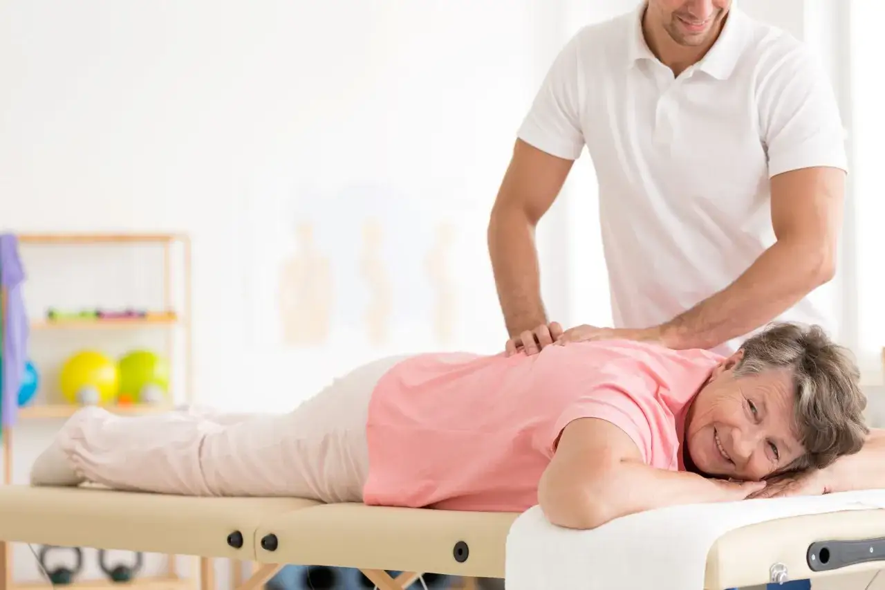 Fizjoterapeuta masuje plecy starszej pani. W tle widać sprzęt rehabilitacyjny, w tym piłki do ćwiczeń. To miejsce, gdzie można skorzystać z zabiegów fango.