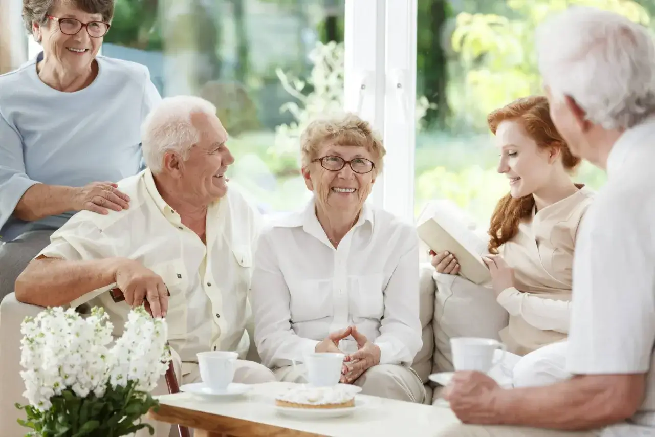 Grupa seniorów i młoda kobieta cieszą się wspólnym, holistycznym popołudniem przy kawie i ciastku.