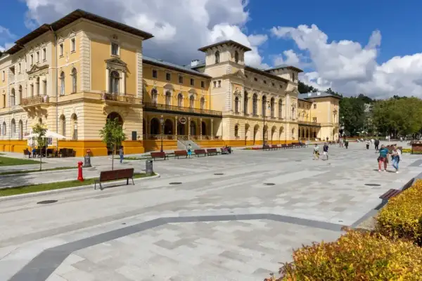 Elegancki budynek sanatorium, plac z ławkami i spacerujący ludzie. Dowiedz się o nowe zasady wyjazdu do sanatorium na NFZ.