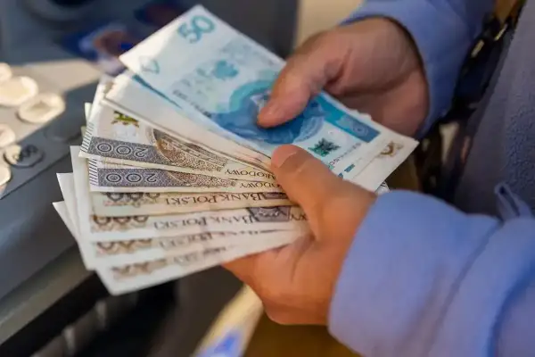Dłonie trzymają stos polskich banknotów, symbolizujących waloryzację emerytury. Widać nominały 50, 100 i 200 zł.
