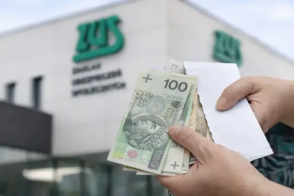 Ręce trzymają polskie banknoty przed budynkiem ZUS. Wypłata 14 emerytury wreszcie nadeszła.