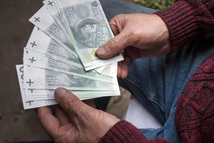 Dłonie seniora trzymają stos banknotów 100 zł. To może być pomoc z programu 800 plus dla seniorów.