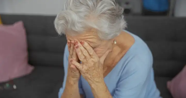 Starsza kobieta z siwymi włosami zakrywa twarz dłońmi, być może doświadczając trudnych emocji związanych z objawami alzheimera.